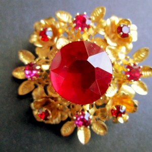 Peut inclure: Broche florale dorée avec une grosse pierre rouge facettée au centre. De plus petites pierres rouges et roses sont serties dans les pétales. La broche présente un design vintage détaillé.