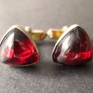 May include: A pair of cufflinks featuring triangular, deep red glass stones set in a silver-coloured metal frame. The cufflinks have a gold-coloured metal backing. The background is a dark grey colour.