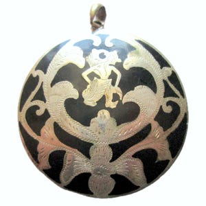 以下が含まれることがあります： 精巧な銀色のフィリグリーと中央の人物が特徴の、丸い黒いペンダント。ペンダントの上部には、吊り下げ用の銀色のループが付いています。デザインは、花柄と様式化された人物が特徴です。