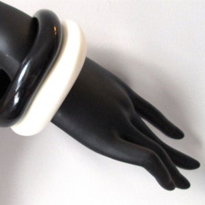 May include: Two bangle bracelets, one black and one white, worn on a mannequin's wrist.