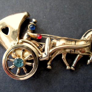 May include: Gold-tone brooch depicting a horse-drawn carriage with a driver. The carriage has a heart-shaped window and a green jewel in the wheel. The horse is detailed with black accents.