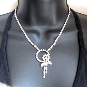 May include: A silver rhinestone necklace with a dangling flower design. The necklace is on a black mannequin.