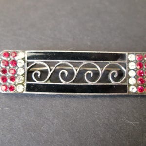 May include: A silver bar brooch with a black central panel and decorative scrollwork. The ends have a geometric design with red and clear rhinestones. A vintage jewellery item.