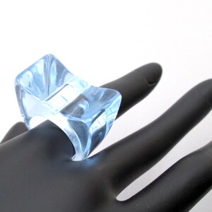 May include: A light blue, clear, plastic ring with a unique, geometric design. The ring is worn on a model's finger.
