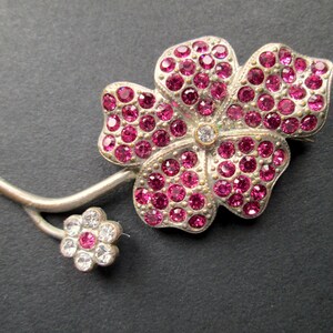 May include: A silver brooch in the shape of a flower with pink rhinestones. The flower has five petals and a single clear rhinestone in the centre. The brooch has a stem with a smaller flower with clear rhinestones.