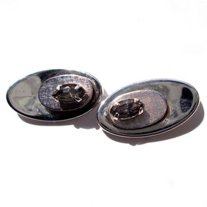 May include: A pair of silver cufflinks with a clear rhinestone in the centre of each. The cufflinks are oval shaped and have a textured surface.