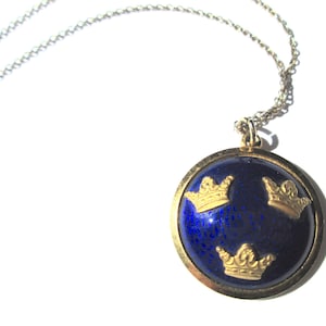 May include: A silver chain necklace with a round pendant. The pendant has a gold-colored rim and a dark blue center with three gold-colored crown designs. The necklace is displayed on a white background.