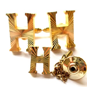 May include: A set of gold-tone cufflinks and a tie tack featuring the letter "H" in a textured, sunburst design. The tie tack has a chain and a round back.