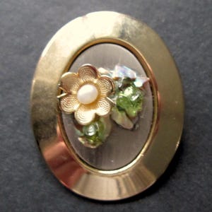 May include: Oval gold-tone brooch with a floral design. The brooch features a gold flower with a pearl centre and green crystal accents. The brooch is approximately 3.8 cm in length.