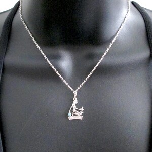 May include: A silver necklace with a pendant depicting a seated figure holding a cross and a small turquoise stone. The necklace is shown on a black mannequin.