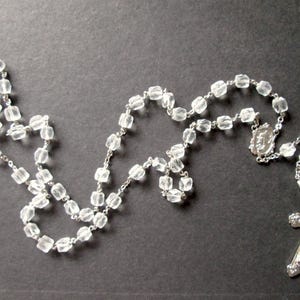May include: A rosary featuring clear, faceted, cube-shaped beads linked by a silver-toned metal chain. The piece includes a central silver-toned metal centrepiece depicting a figure and terminates in a silver-toned metal crucifix with rounded ends.