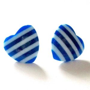 May include: A pair of heart-shaped earrings. Each earring is blue with white diagonal stripes. The earrings are set against a white background, with shadows visible below them.