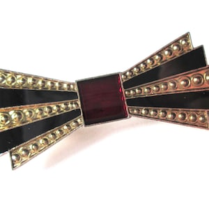 May include: A gold and black bow brooch with a red centre. The bow is made of metal and has a textured surface.