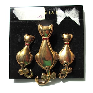 May include: A set of three gold-tone cat brooch pins with red rhinestone accents. The cats are facing forward with their tails curled up behind them.