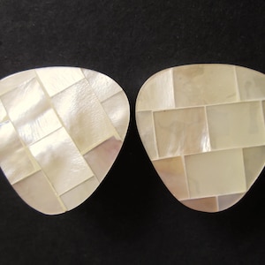 May include: Two mother-of-pearl guitar picks with a mosaic design. The picks have a light, iridescent sheen. The mosaic pattern is made up of small, rectangular pieces. The picks are on a dark background.