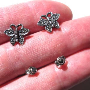 May include: A pair of silver butterfly earrings with black marcasite stones. The earrings are shown on a hand.