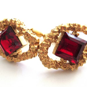 May include: A pair of gold cufflinks with red square-shaped gemstones set in a textured gold frame.