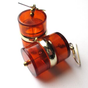 Peut inclure: Une paire de boucles d'oreilles de couleur ambre avec des accents dorés. Les boucles d'oreilles sont en forme de petits barils avec une bande dorée autour du milieu.