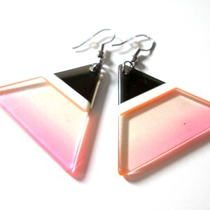 May include: A pair of geometric earrings with a black, white and pink ombre design. The earrings are made of acrylic and have a triangle shape.