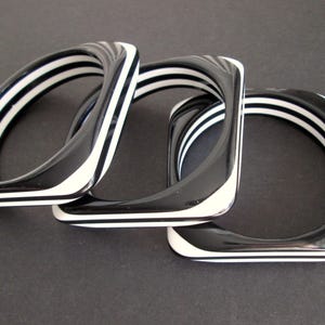 May include: Three square bangles with a black and white striped design. The bangles are made of a glossy material and are arranged in a slightly overlapping pattern against a dark background.