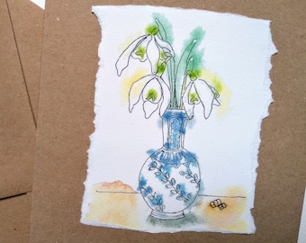 May include: Watercolor painting of snowdrop flowers in a blue and white vase. The vase has a detailed floral design. The artwork is on textured paper with a torn edge, set against a brown card. The flowers have white petals and green accents.