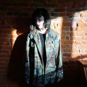 May include: A person wearing a camouflage jacket with a black shirt underneath. The person is standing in front of a brick wall.