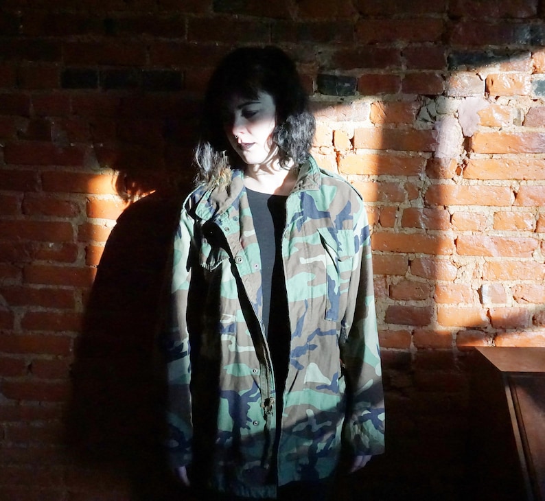 May include: A person wearing a camouflage jacket with a black shirt underneath. The person is standing in front of a brick wall.