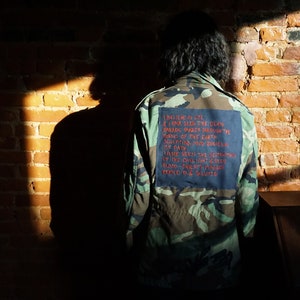 May include: A camouflage jacket with a blue patch on the back. The patch has red text that reads: "I believe in life I have seen the idea invade barren through the torso of the earth molding mud bodies into path I have seen the strength of the daylight between blood thirsty hands pressed to & saluted"