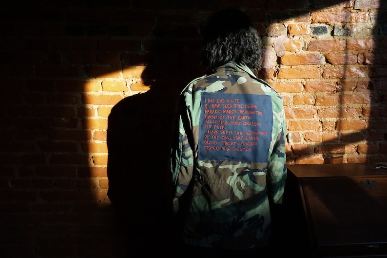 May include: A camouflage jacket with a blue patch on the back. The patch has red text that reads: "I believe in life I have seen the idea invade barren through the torso of the earth molding mud bodies into path I have seen the strength of the daylight between blood thirsty hands pressed to & saluted"