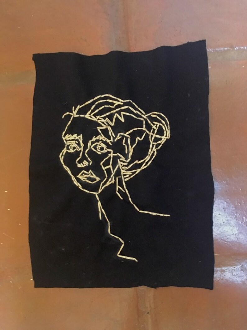May include: A black fabric patch with a gold embroidered outline of a person's face and hair.