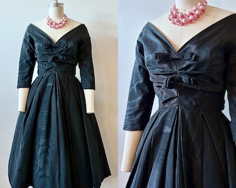 Può includere: Un abito vintage nero con scollatura a V e maniche a tre quarti. L'abito presenta un corpetto aderente con dettagli a fiocco e una gonna ampia e plissettata. Una collana di perline rosa è drappeggiata attorno al collo del manichino.