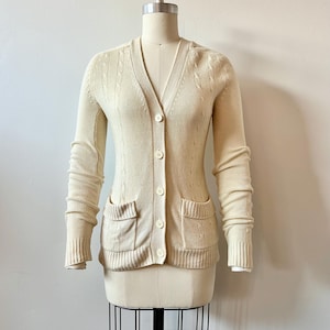 Puede incluir: Un cárdigan de punto de color crema con un patrón de punto de cable, mangas largas y cierre de botones en la parte delantera. Tiene dos bolsillos de parche en la parte delantera y un cuello en V.