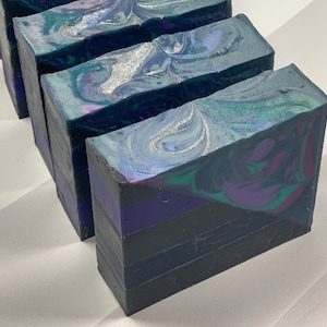 May include: Four rectangular bars of handmade soap. The top of each bar has a swirling pattern of teal, silver, and purple. The bottom of each bar has horizontal stripes of black, purple, and navy blue.