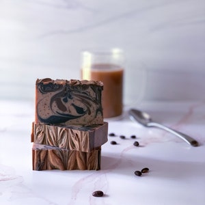 May include: Three bars of handmade coffee soap stacked on top of each other on a white marble surface. The soap is brown and black with swirls of color. There are coffee beans scattered around the soap.