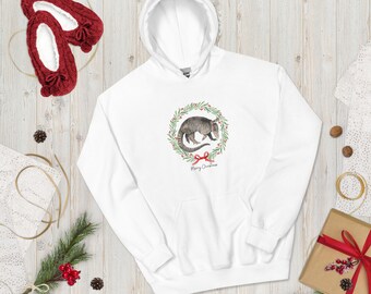 May include: White hoodie featuring a watercolor illustration of an armadillo inside a Christmas wreath with a red bow and the words "Merry Christmas." The hoodie is surrounded by holiday decorations, including red slippers.