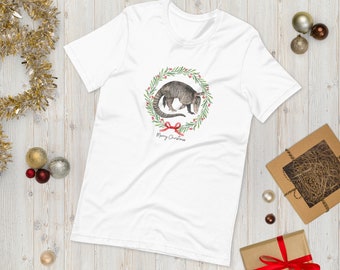 May include: White t-shirt featuring a watercolor illustration of an armadillo inside a Christmas wreath with red berries and a red bow. The words "Merry Christmas" are written below the wreath. The shirt is a short-sleeved crew neck.