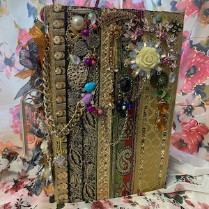 May include: A gold and black journal adorned with a variety of embellishments, including beads, chains, ribbons, and rhinestones. The journal is decorated with a white rose and other floral accents.