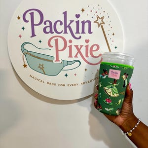 May include: A green neoprene cup sleeve featuring a repeating pattern of animated characters, including a woman in a green dress, a frog, and water lilies. The sleeve has a small rectangular pink label with the text Packin Pixie printed in black.