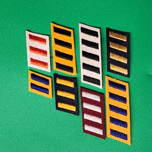 May include: A collection of seven rectangular embroidered patches featuring various color combinations. The patches display horizontal stripes in red, yellow, white, black, blue, and maroon, arranged in different patterns across the fabric pieces.