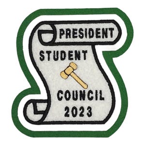 Puede incluir: Un parche de fieltro blanco y verde con un diseño de rollo. El rollo dice "Presidente Consejo Estudiantil 2023" y tiene una imagen de martillo.