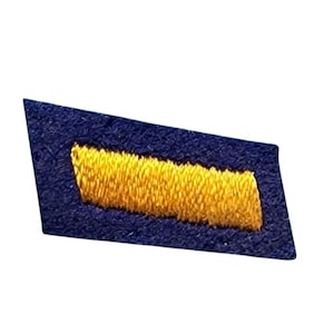 May include: A trapezoidal-shaped fabric patch featuring a dark blue base with a single, thick, horizontal yellow embroidered bar centered across the middle. The embroidery has a textured, raised appearance against the flat blue fabric background.