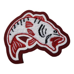 May include: Embroidered patch in the shape of a fish, featuring a white body with red and black details. The fish is outlined in white and set against a maroon background. This patch is suitable for jackets, bags, or other fabric items.
