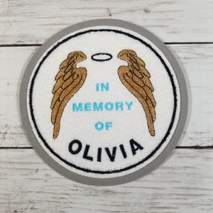 Può includere: Una toppa rotonda bianca con ali d'angelo dorate e un alone. Il testo "IN MEMORY OF OLIVIA" è ricamato in verde acqua e nero. La toppa ha un bordo grigio e un contorno nero.