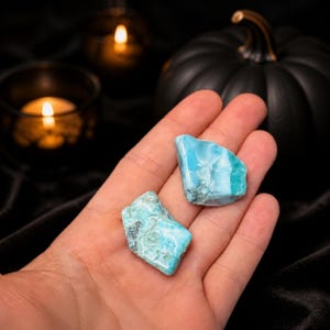 May include: Two polished, irregular pieces of light blue and white Larimar gemstone with visible marbling and crystalline textures. The stones display a vibrant sky-blue hue with cloudy white patterns, showcasing a smooth, natural, and organic shape.