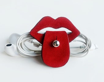 May include: Red leather cord organiser shaped like a pair of lips with a tongue sticking out. The organiser is holding a pair of white earbuds.