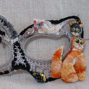 May include: A black and silver masquerade mask with a variety of embellishments, including cats, butterflies, and beads. The mask is made of lace and fabric.