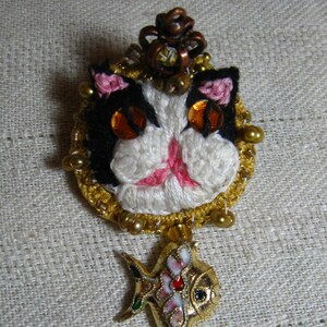 May include: A gold and white crocheted cat brooch with a crown and a dangling gold fish charm with a white and pink enamel design.