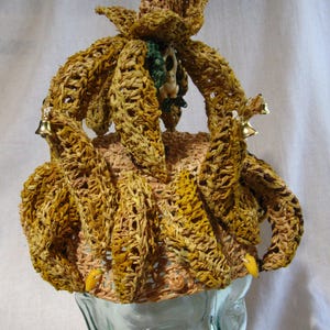 May include: A crocheted hat made of straw, shaped like a banana bunch with a small, pointed top. The hat is decorated with small bells and a small, white, crocheted figure.