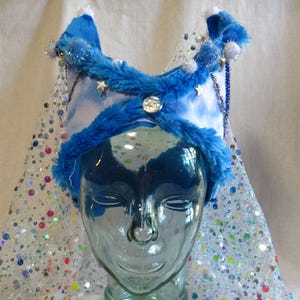 Puede incluir: Una corona de fantasía azul y blanca con un ribete azul esponjoso, estrellas plateadas y un velo transparente con lentejuelas. La corona está adornada con detalles plateados y un botón decorativo grande. El velo está decorado con lentejuelas de colores y pequeñas cuentas transparentes.