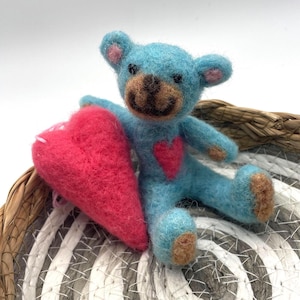 Könnte beinhalten: Ein handgefertigter blauer Teddybär mit brauner Schnauze und einem roten Herz auf der Brust, der ein großes rotes Herz hält. Der Bär ist aus Filz und sitzt auf einem geflochtenen Korb. Ein bezaubernder, handgefertigter Artikel.